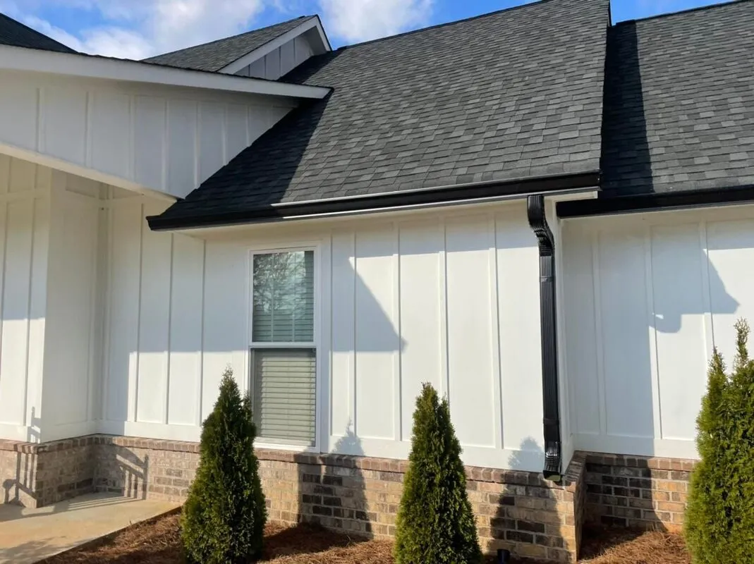 Black gutter and downspout along the roofline of a white board-and-batten house with a brick base