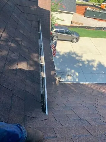 Rooftop view down a roof valley with the gutter packed full of dark debris, a worker's boot in the foreground and a West Georgia driveway with a parked SUV below