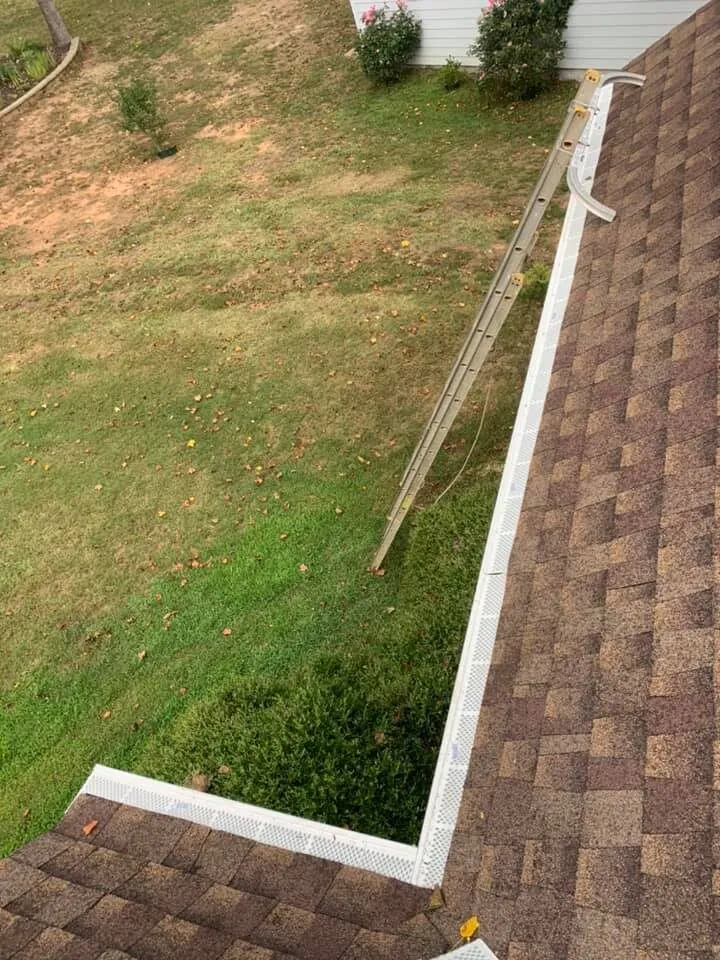View down a shingle roof edge with white gutter guard seated along the run and around the corner, extension ladder at the eave, lawn below