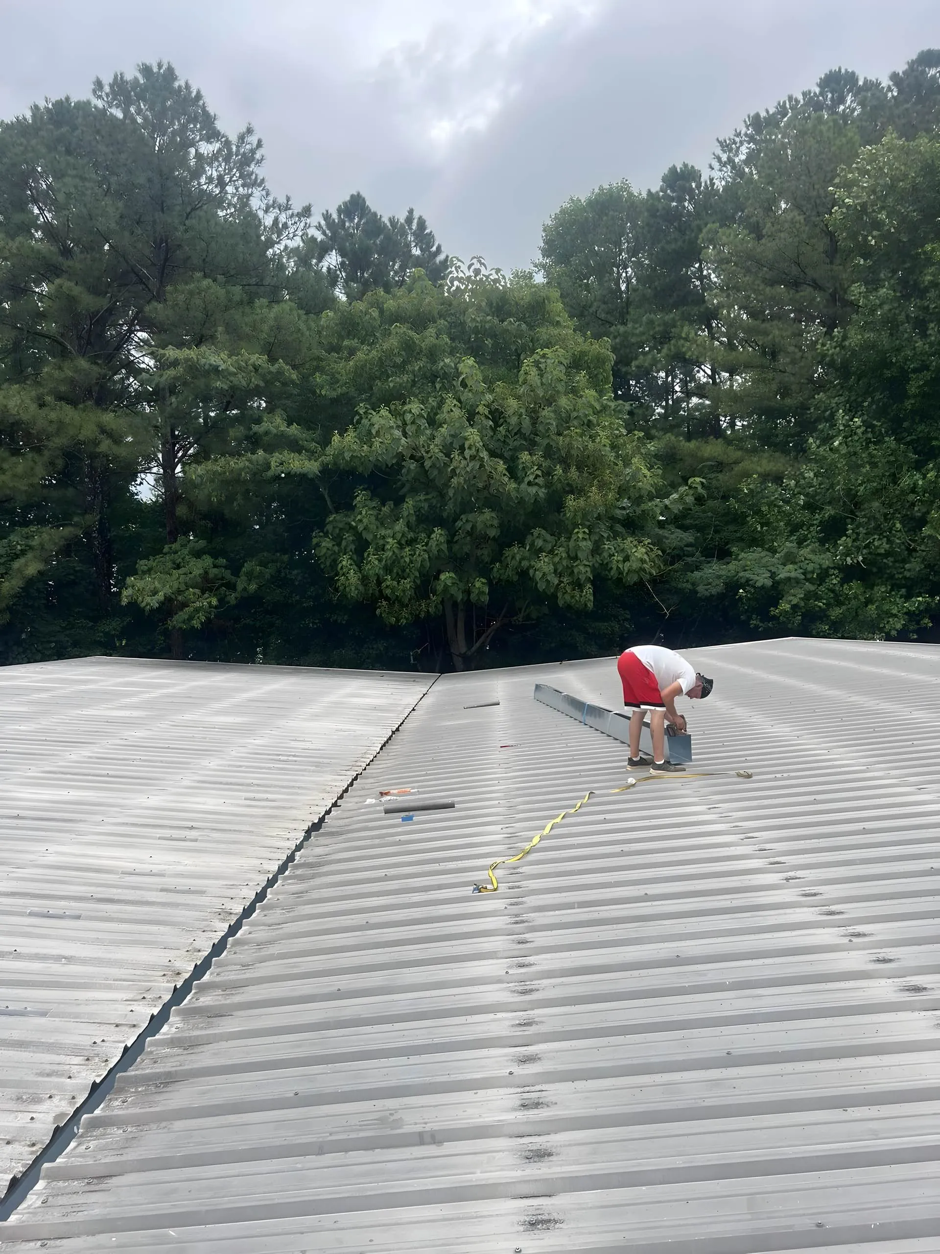 Crew member fastening a seamless gutter section on a wide metal roof, pine trees behind