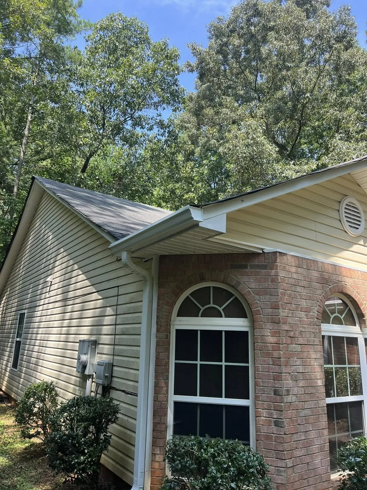 New white seamless gutter and downspout on the gable corner of a cream ranch with brick arch windows, heavy oak and pine canopy above