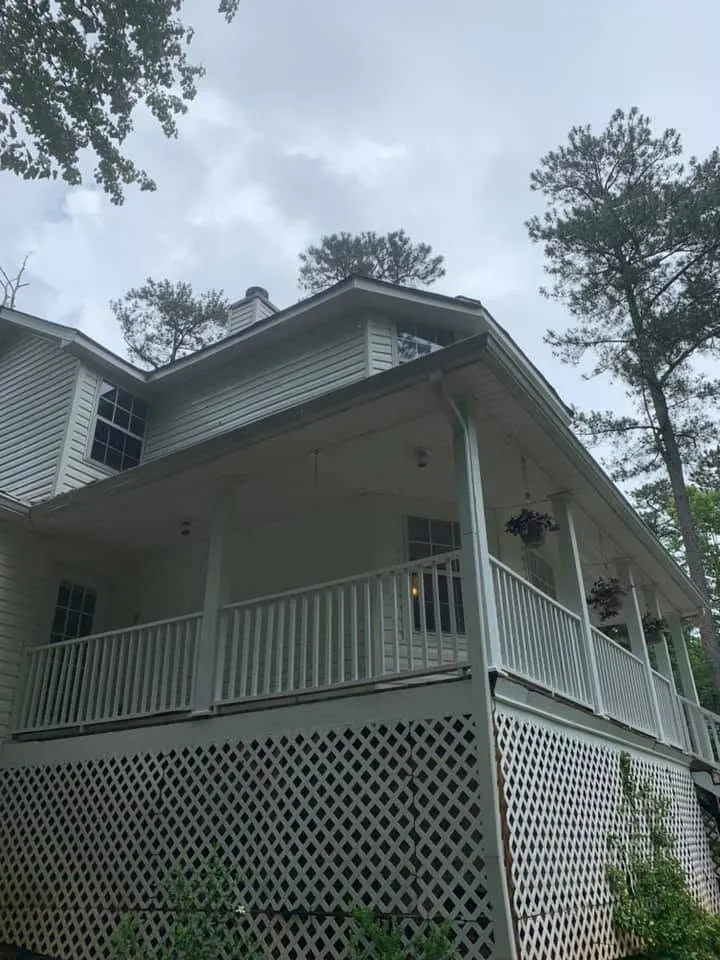 White two-story farmhouse porch corner with lattice skirting and chimney, clean siding after a house wash, pines behind