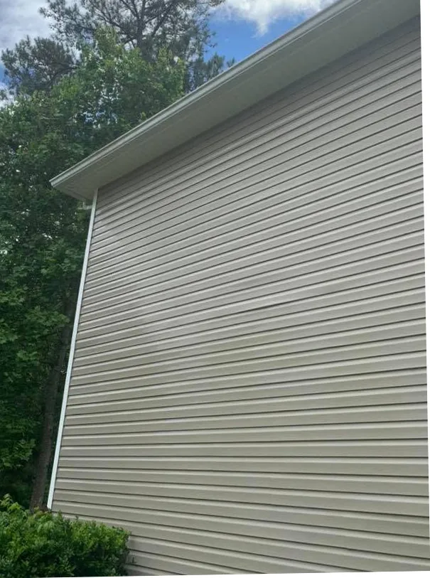 Clean tan vinyl siding gable wall under a white gutter and blue sky, green trees beside it, after a house wash