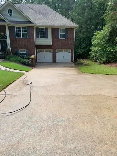 Long, freshly pressure-washed concrete driveway leading to a brick two-story home with a double garage, hose still laid across the drive