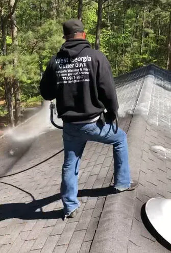 Technician in a branded West Georgia Gutter Guys hoodie spraying a soft wash across a shingle roof ridge