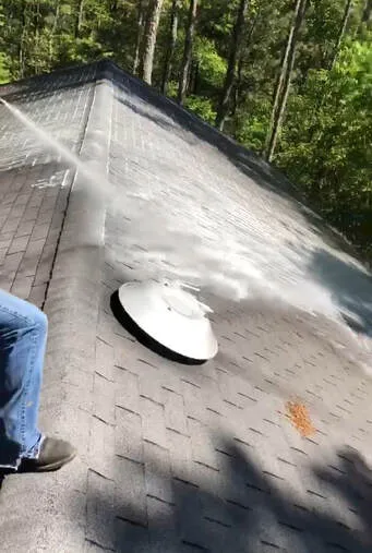 Soft wash spray drifting over a shingle roof and vent cap, technician at the edge of the frame