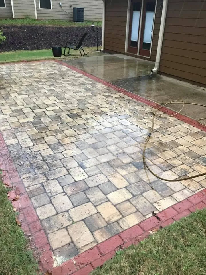 Paver patio with dark mottled staining across the blocks, garden hose lying over the pavers, red brick border