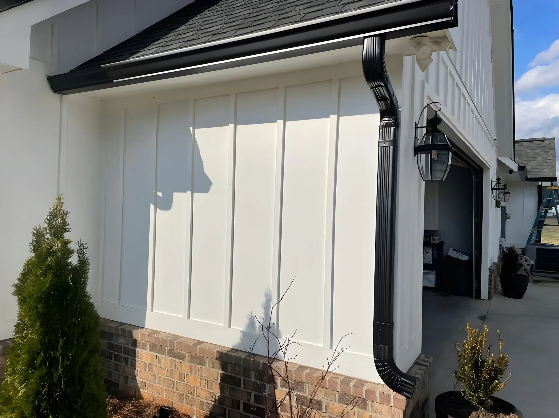 New black seamless gutter and downspout on a white board-and-batten farmhouse wall above a brick foundation